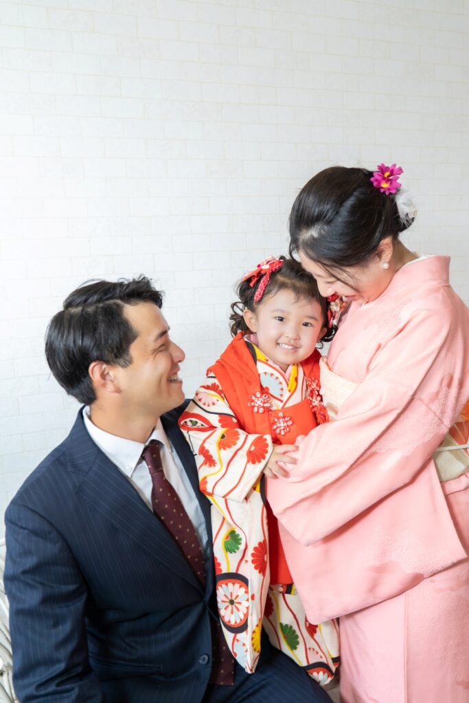 おうちスタジオの白いレンガ調の壁の前で、七五三のお祝いをする3人家族。中央には華やかなアンティーク風の花柄着物を着た3歳の女の子が笑顔でカメラを見ている。隣にはピンク色の訪問着を着用し、フューシャピンクの花の髪飾りを付けたお母さんが女の子を見つめ、ダークネイビーのスーツを着たお父さんも笑顔で女の子を見ている。幸せそうな瞬間を捉えた家族フォト